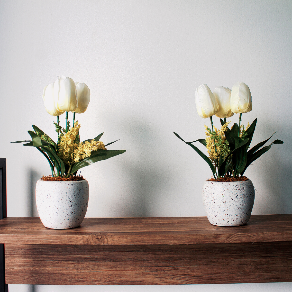 Two white plant pots on a wooden table. Each pot has white tulips blooming in it with long green leaves.